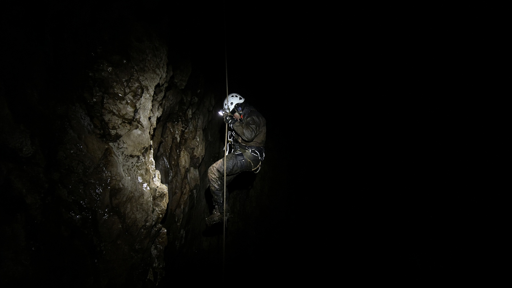 A caver on rope descending into deep darkness, lit by a single burst of flash.