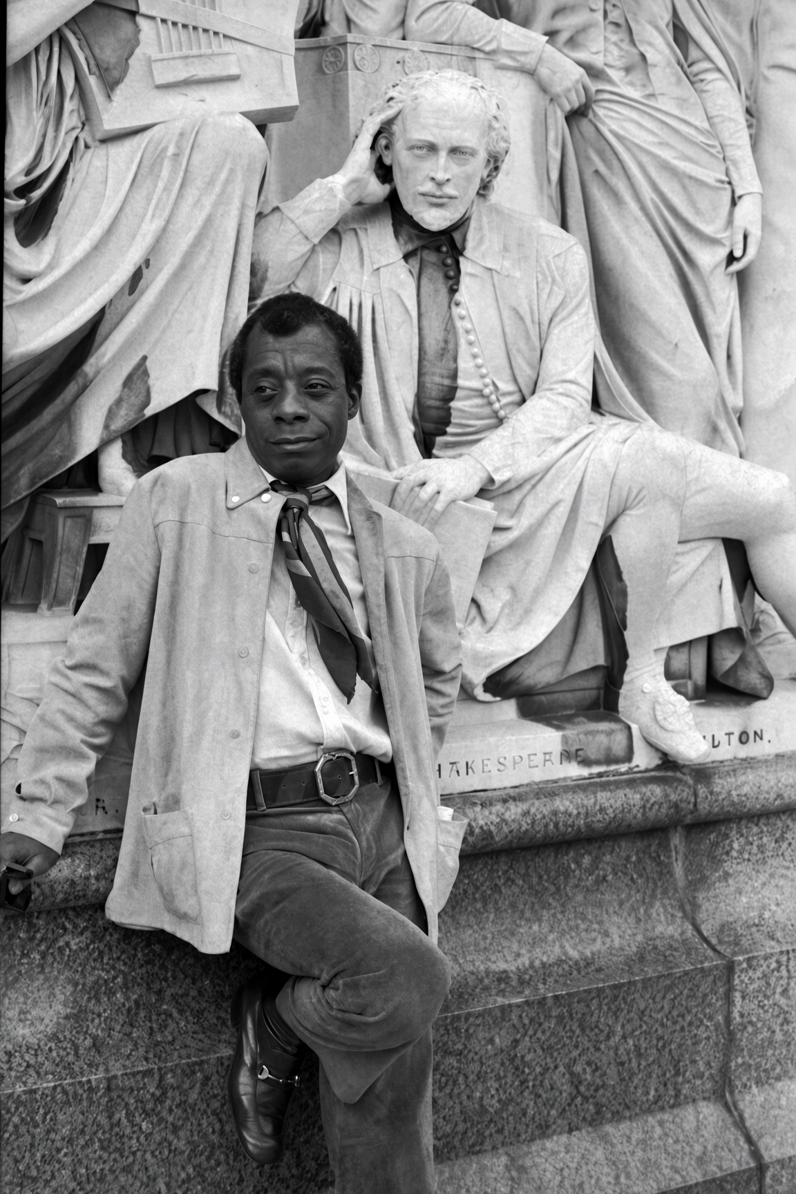 James Baldwin seated against a stone monument, looking directly at the camera.