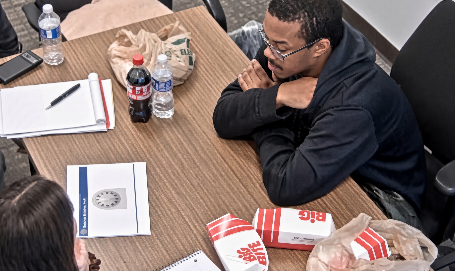 Interrogation-room still: a young man in a dark hooded sweatshirt seated at a conference table with a legal pad, a diagram, bottled drinks and fast food, hands folded near his face